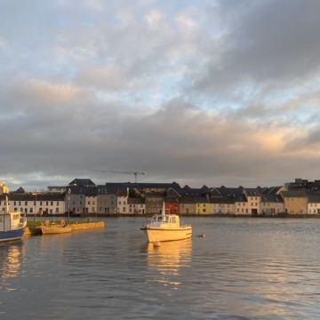 A serene sunset overlooks the Long Walk in County Galway, Ireland, with vibrant orange and pink hues reflecting on the calm waters. Historic colorful houses line the waterfront promenade.